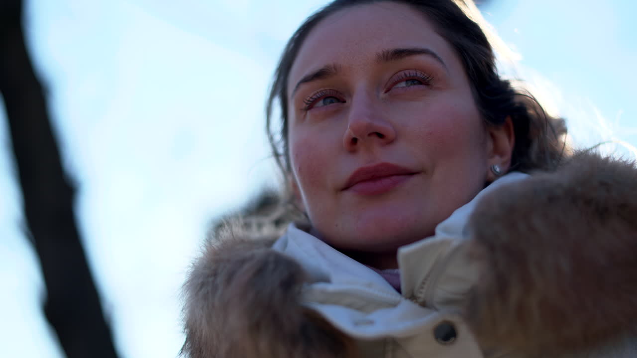A woman wearing a white coat with fur collar, walking outside on a bright winter day with sunlight flare