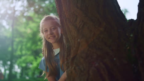 Cute-blond-girl-looking-out-huge-tree-trunk.-Child-touching-bark-on-sunny-day.