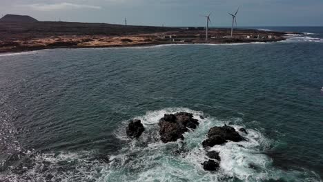 Espuma-De-Mar-Que-Rodea-Piedras-Y-Molinos-De-Viento-En-La-Costa-De-La-Playa-De-Corralejos,-Fuerteventura