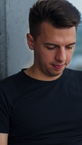 dark-haired young man focused on his work at laptop. male sitting near the panoramic window in a comfortable chair. blurred backdrop. vertical video