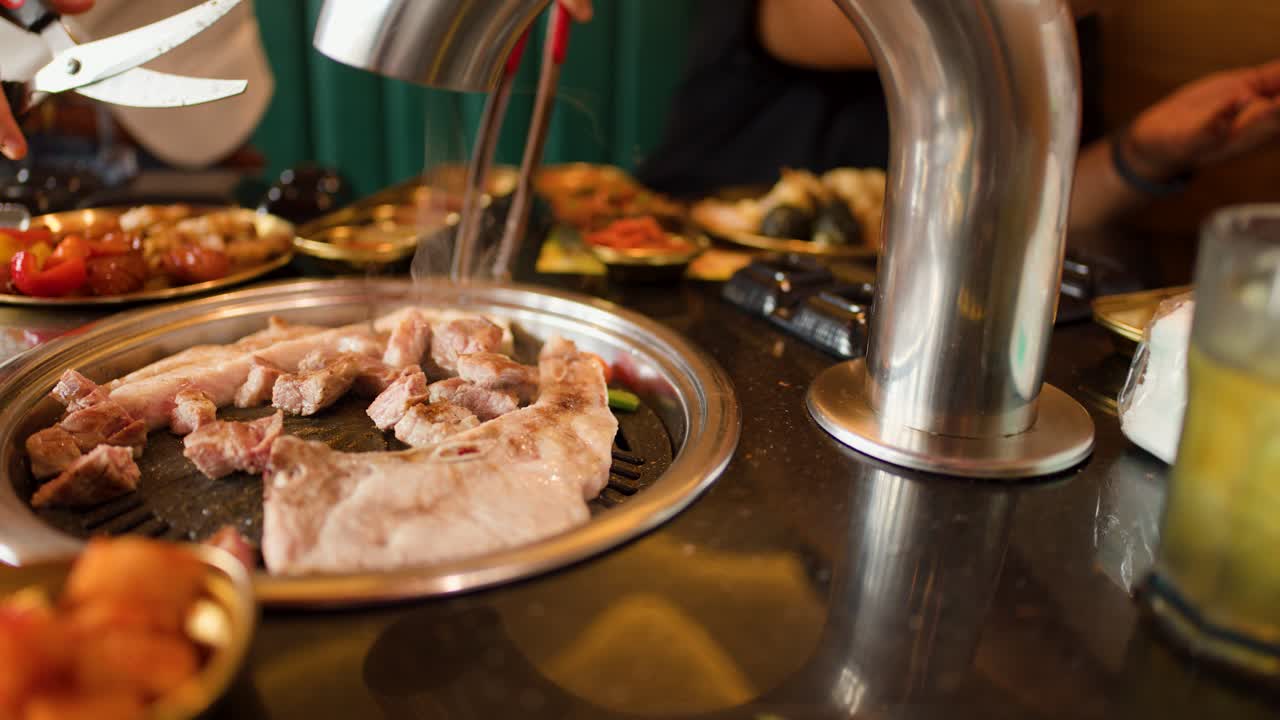 Hands grill pork and mushrooms on tabletop grill, surrounded by side dishes, warm lighting, close-up