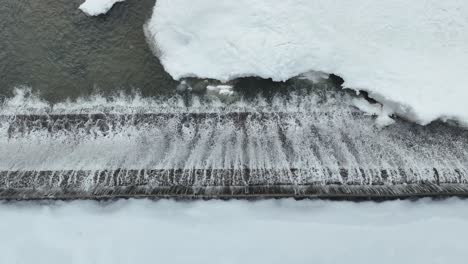 a top-down drone shot capturing the water cascading over a frozen dam, with ice and snow covering the landscape. the drone moves from right to left, showing the stunning winter scene.