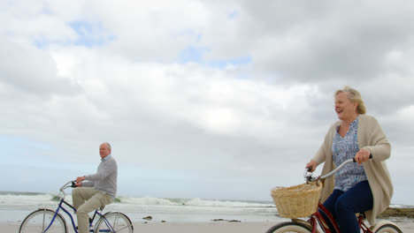 Vista-Lateral-De-Una-Pareja-De-Ancianos-Caucásicos-Montando-Bicicleta-En-La-Playa-4k