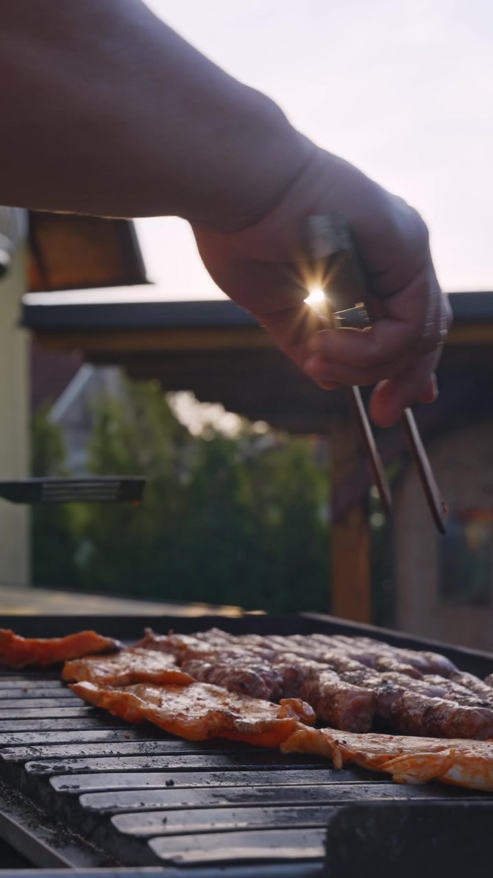 Succulente fette di carne sfrigolano su una griglia per barbecue, una mano le gira con le pinze in un accogliente cortile. Video verticale, tramonto caldo, perfetto per storie social.