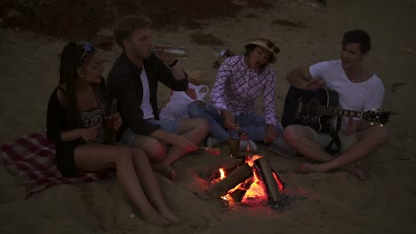 group of young and cheerful people sitting by the fire on the beach in the evening, grilling sausages and playing guitar