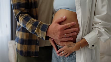 Close-up-of-a-happy-man-in-a-plaid-shirt-stroking-his-wife's-pregnant-belly-at-home-while-touching-her-hand