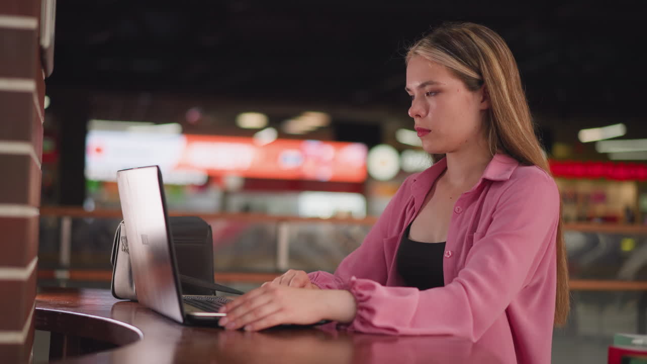 dama de vestido rosa sentada en un centro comercial con su bolsa negra en la mesa de madera, trabajando en su portátil, el fondo está ligeramente borroso con personas moviéndose, con efectos de luz bokeh