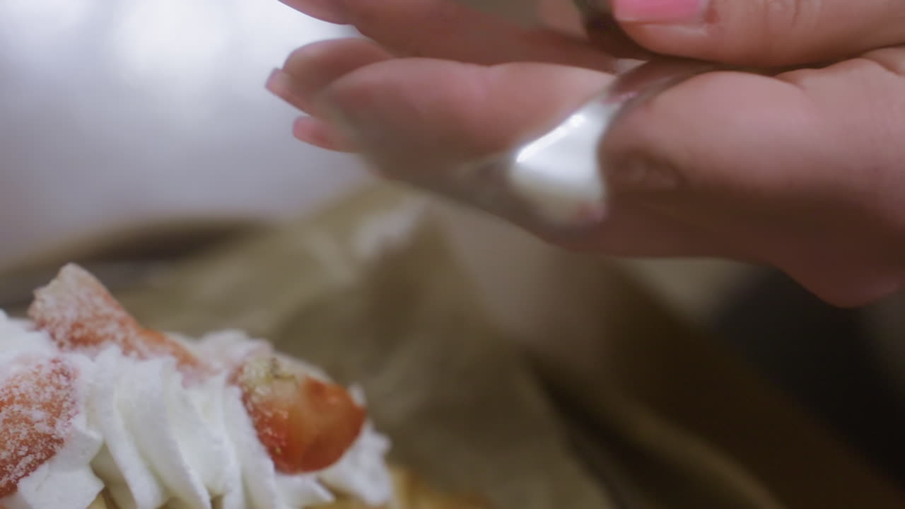 Close-up of female hand unwrapping fork with napkin in cozy cafe, croissant with whipped cream and coffee cup on table. Soft lighting creates relaxed ambiance in background