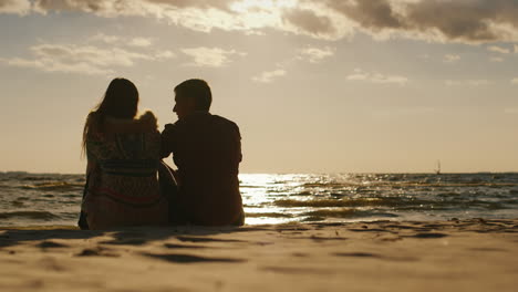 Siluetas-De-Parejas-De-Enamorados-Sentados-En-La-Playa-Mirando-El-Atardecer-Y-Un-Tablero-Con-Una-Vela-De-Regreso