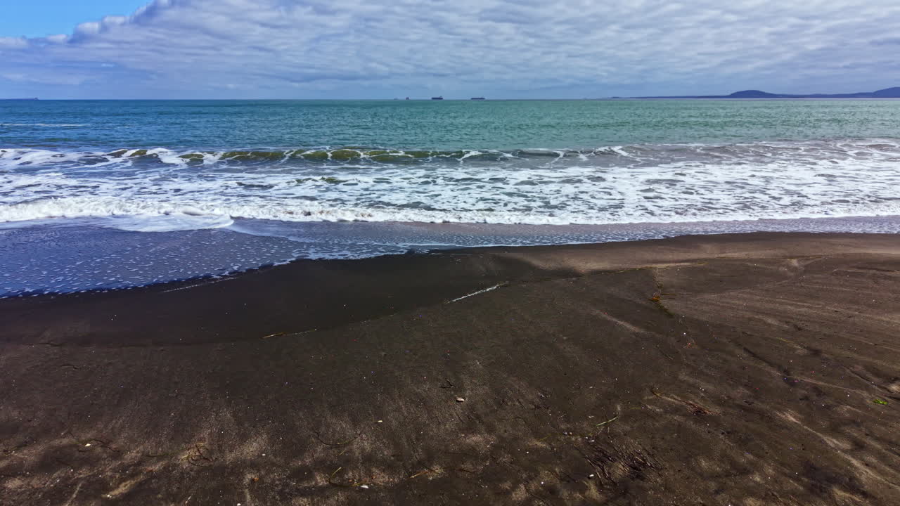 Le onde si infrangono su una spiaggia di sabbia scura con cieli nuvolosi sull'oceano a mezzogiorno.