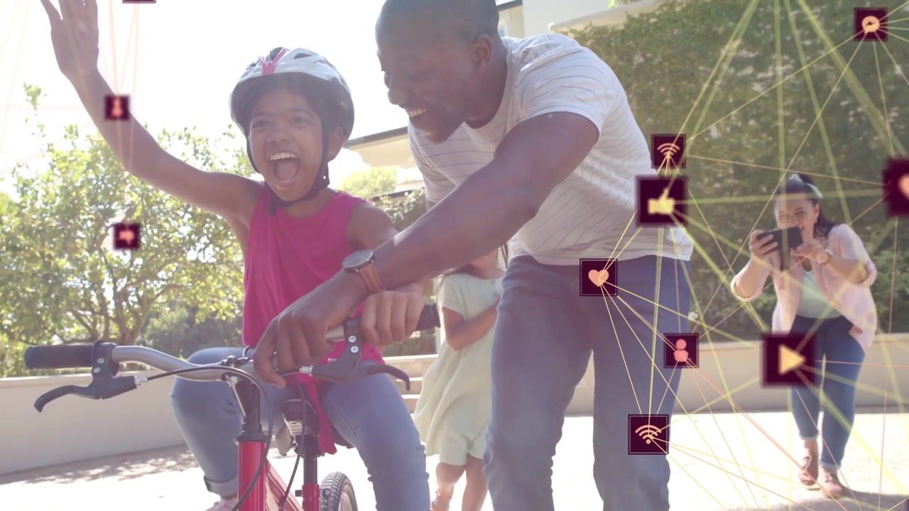 Man guiding girl learning on red bike, easing grip while tech icons overlaying, mom filming phone