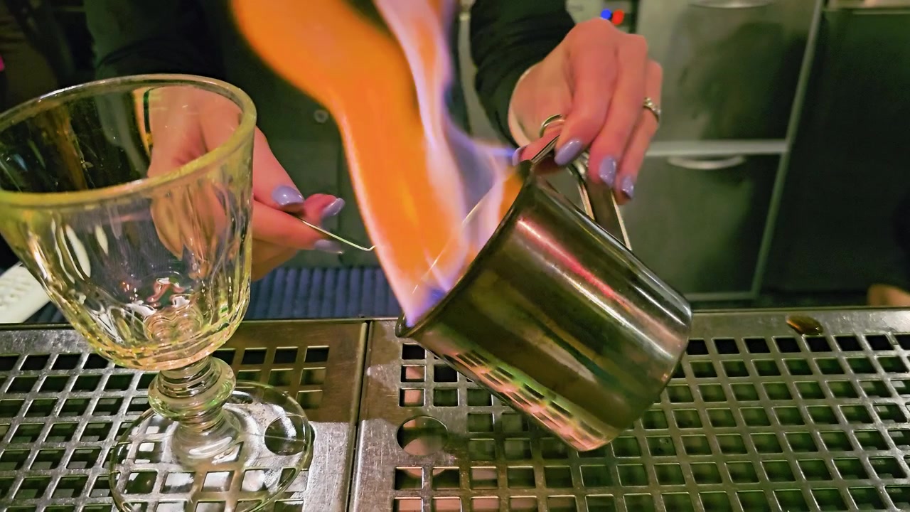Primo piano atmosferico di un bar che mostra un rituale di cocktail fiammeggiante, zolletta di zucchero su cucchiaino forato, tazza di metallo, calice di vetro, mani del barista e postazione per bevande in acciaio inossidabile riflettente