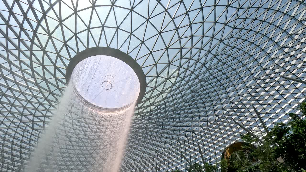 Camera tilts down revealing indoor waterfall, lush greenery, glass dome, and visitors in natural light