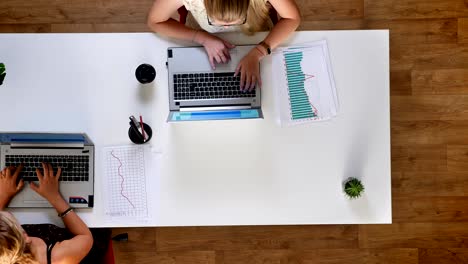 fotografía superior, fotografía en movimiento de dos mujeres sentadas en un escritorio de trabajo sobre un fondo de madera y escribiendo, motivadas, teléfono de pantalla azul usando