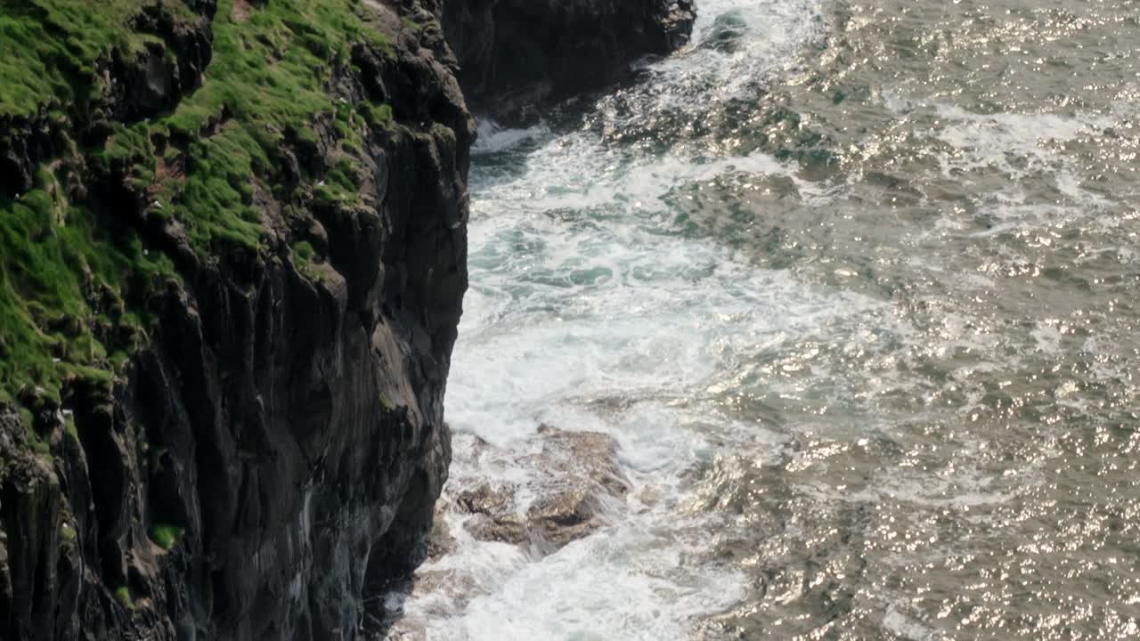 A rocky cliff with green grass overlooks the Faroe Islands’ ocean waves crashing below