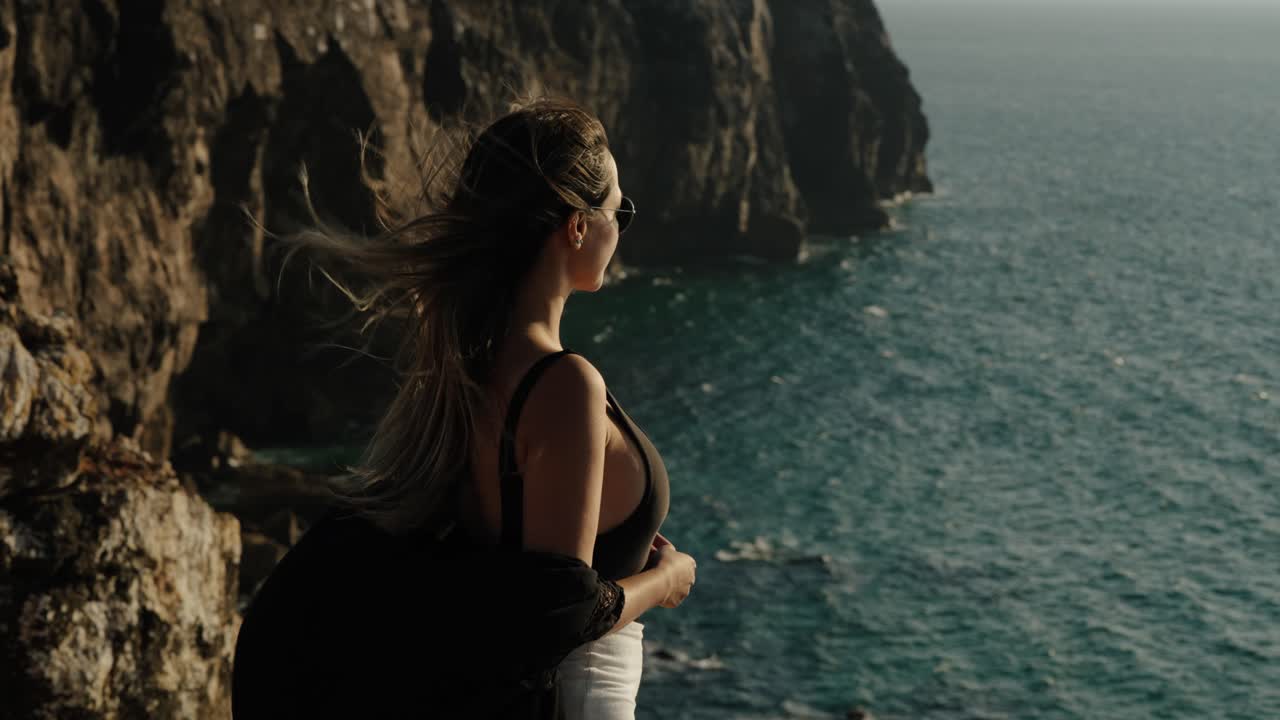 A woman with windswept hair gazes toward the rugged coastline and deep turquoise waters in Algarve, Portugal