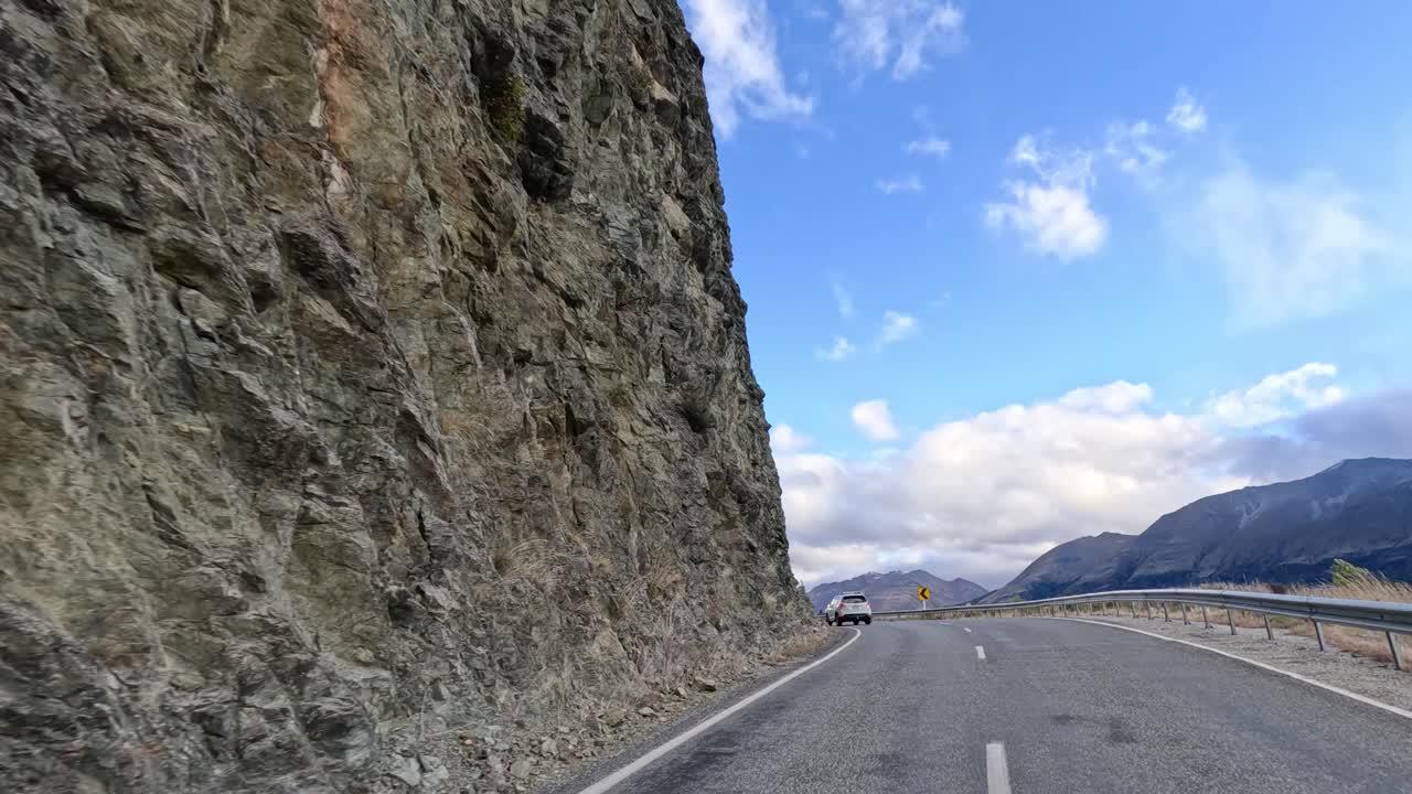 Vehicle travels beside rocky cliff on winding road, daylight, partly cloudy sky, forward camera movement