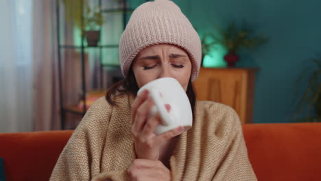 sick young woman wear hat wrapped in plaid sit alone shivering from cold on sofa drinking hot tea