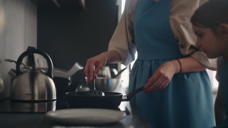 woman is cooking breakfast frying pancakes or eggs in frying pan on stove little daughter is viewing