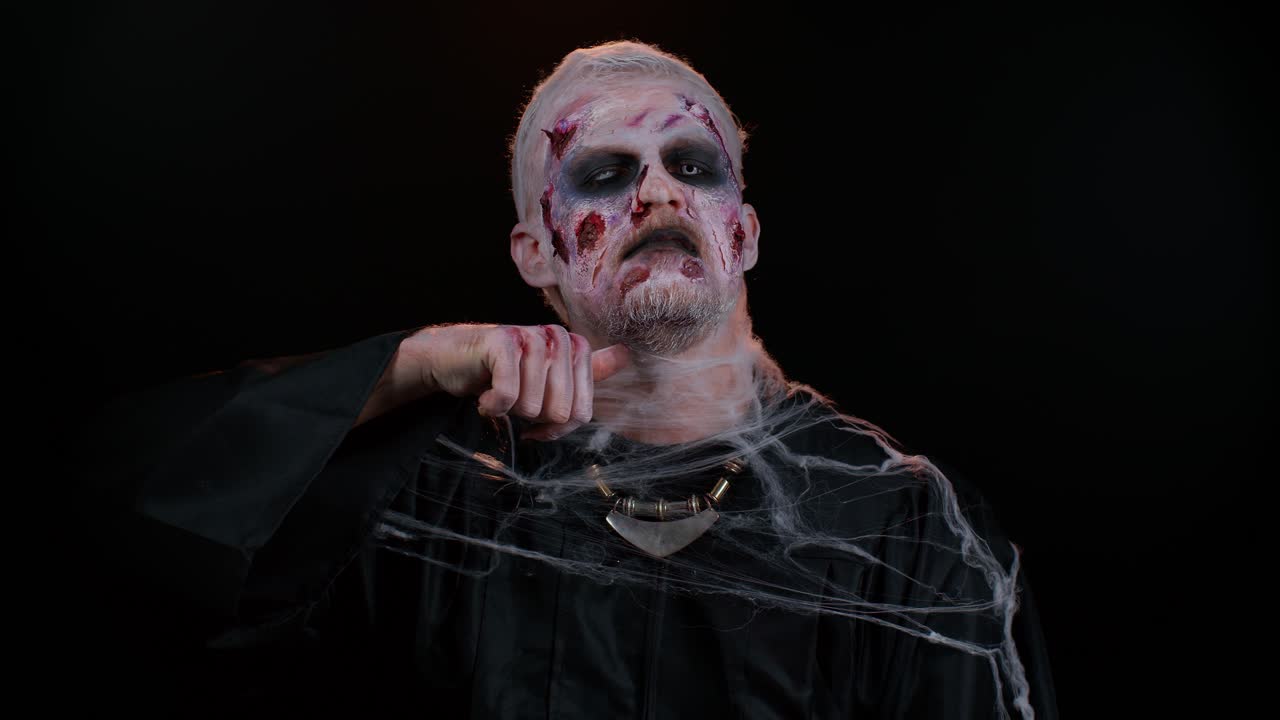 Sinister man Halloween zombie trying to scare showing killing gesture, runs a finger along his neck