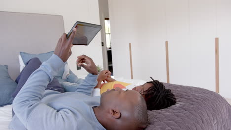 Happy-african-american-father-and-son-lying-on-bed-using-tablet-at-home,-slow-motion