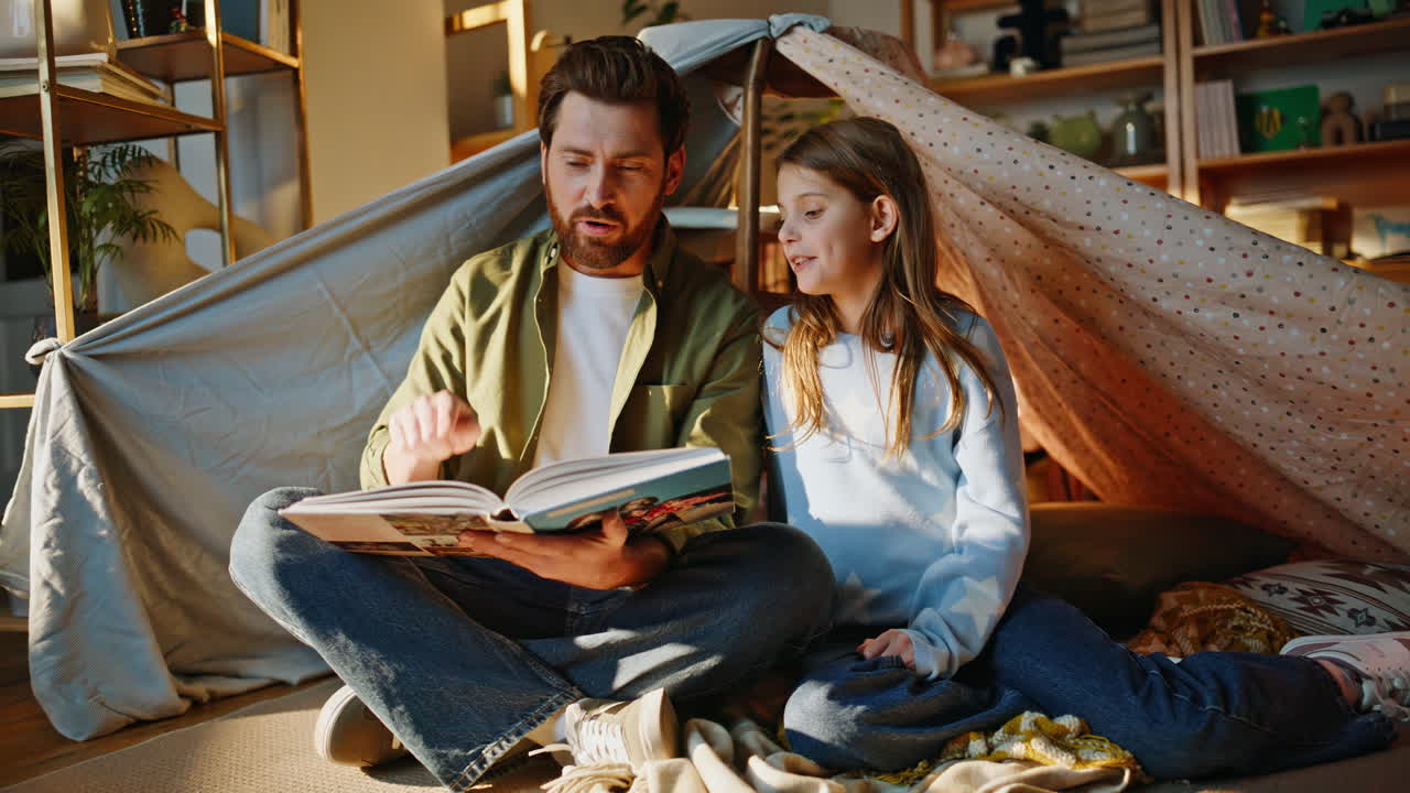 Primer plano de un padre sonriente leyendo un cuento a su hija, arropados con una manta en una cabaña. Vínculo entre padres e hijos