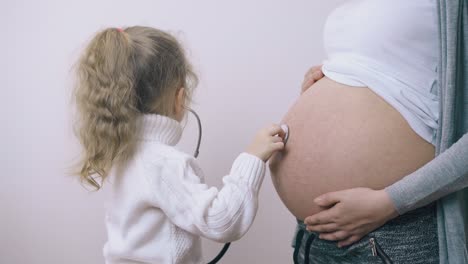 una chica linda le pone un estetoscopio a su madre embarazada con el estómago contra la pared.