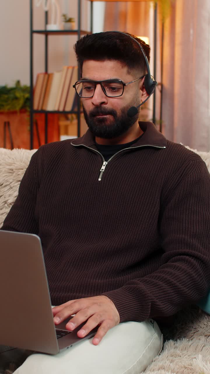 Indian man using wireless headset speaks on laptop video call with client while working at home