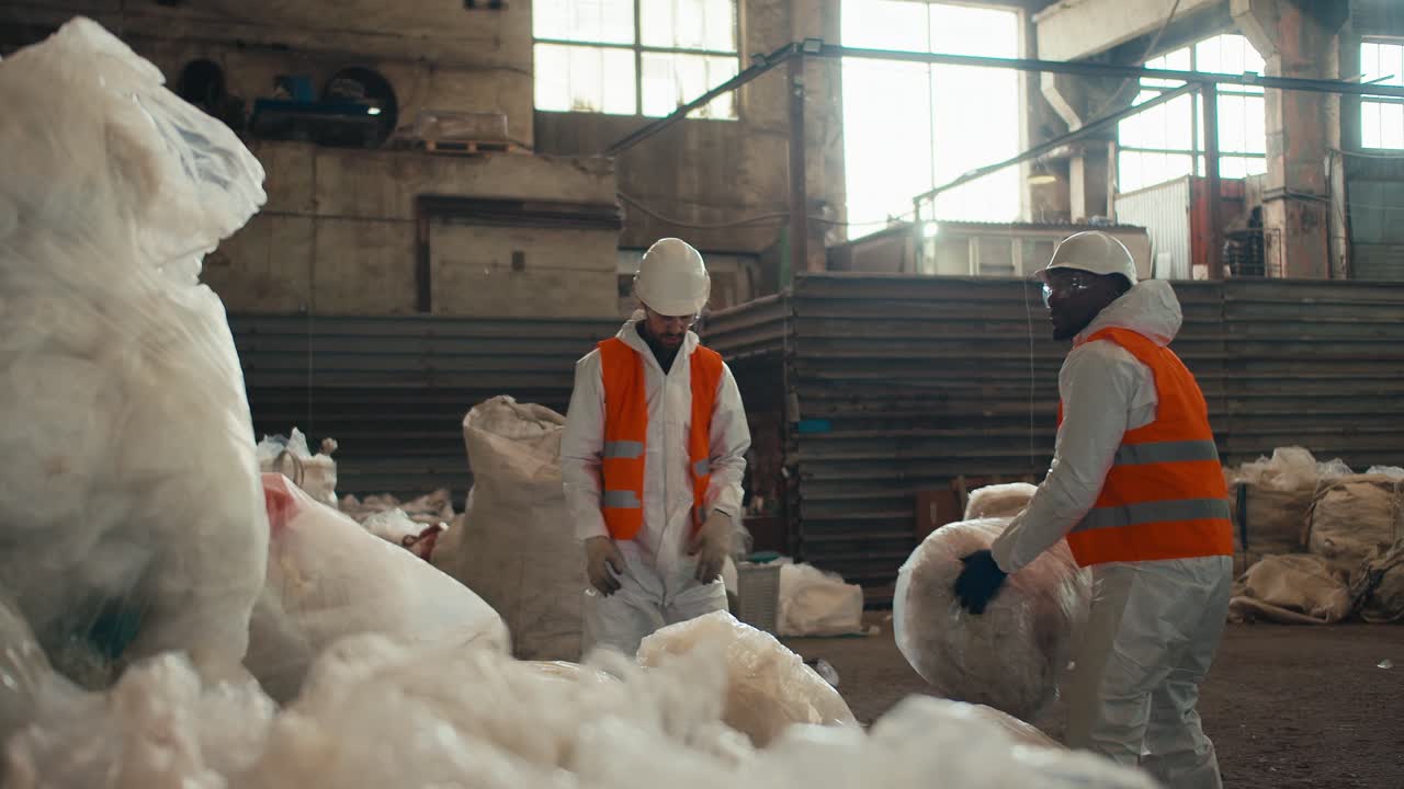 un hombre con piel negra en un uniforme de protección blanco y un chaleco naranja junto con su colega, un hombre con barba apila pilas de celofán reciclado en una pila en una planta de procesamiento y clasificación de residuos