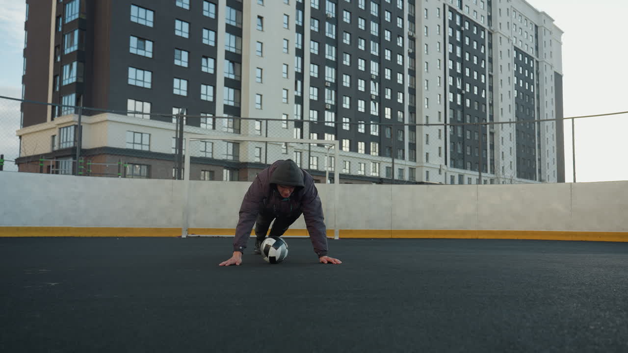 hombre realizando flexiones alternando la colocación de la mano en la pelota de fútbol, mostrando fuerza, equilibrio y aptitud en una arena deportiva al aire libre con postes de gol y edificios residenciales modernos