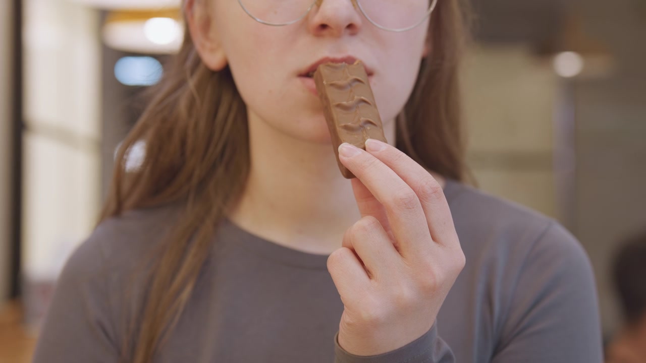 Jovem mulher comendo barra de chocolate lentamente, óculos e suéter neutro, iluminação interna quente, enquadramento macro da boca e dos dedos, provando e saboreando a mordida, fundo de café espontâneo, humor de estudante relaxado