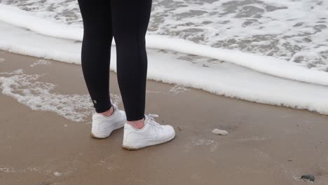 girl in activewear and white trainers stands on the sandy beach as foam from crashing ocean waves flow up to her toes and she runs away on cold grey day