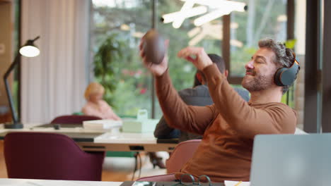 Businessman-With-Feet-On-Desk-In-Office-Listens-To-Music-On-Wireless-Headphones--Catching-Football