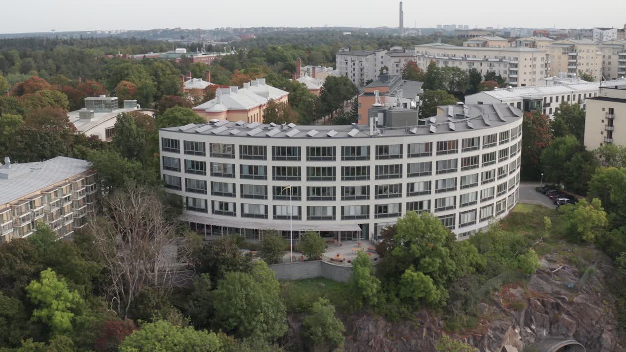 дрон кружит офисное здание с солнечными панелями на вершине с многоквартирными домами и обширным лесом позади в стокгольме, швеция в облачный облачный день