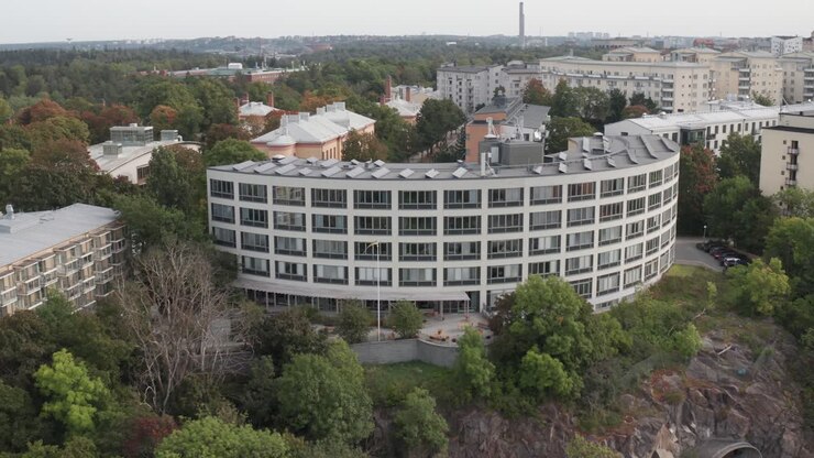 Drone footage circling of office building with solar panels on top with apartment buildings and vast forest behind in Stockholm, Sweden on overcast cloudy day