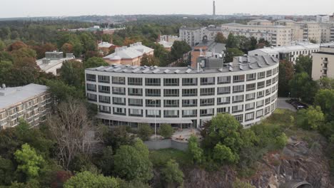 Drone-footage-circling-of-office-building-with-solar-panels-on-top-with-apartment-buildings-and-vast-forest-behind-in-Stockholm,-Sweden-on-overcast-cloudy-day