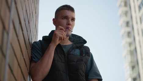 young man wearing dark vest lights cigarette while resting against brick wall with calm expression as tall modern building and faintly visible flying insect appear blurred in background