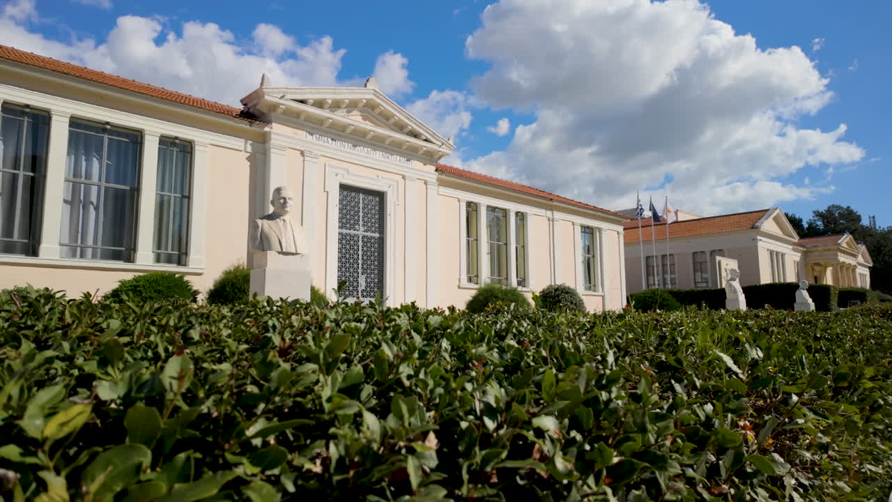 A neoclassical building with busts and manicured hedges in Pafos, Cyprus, under a partly cloudy sky
