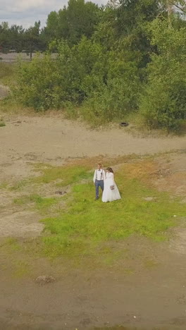 happy newlywed couple walks along bank of calm lake to water edge near green park on autumn day aerial view