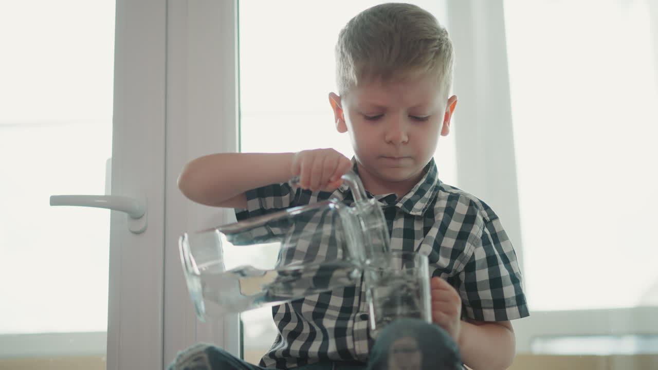 kid sitting by window pouring water from transparent jar into glass cup placed on leg and drinking during bright daylight with relaxed expression wearing checkered shirt and jeans