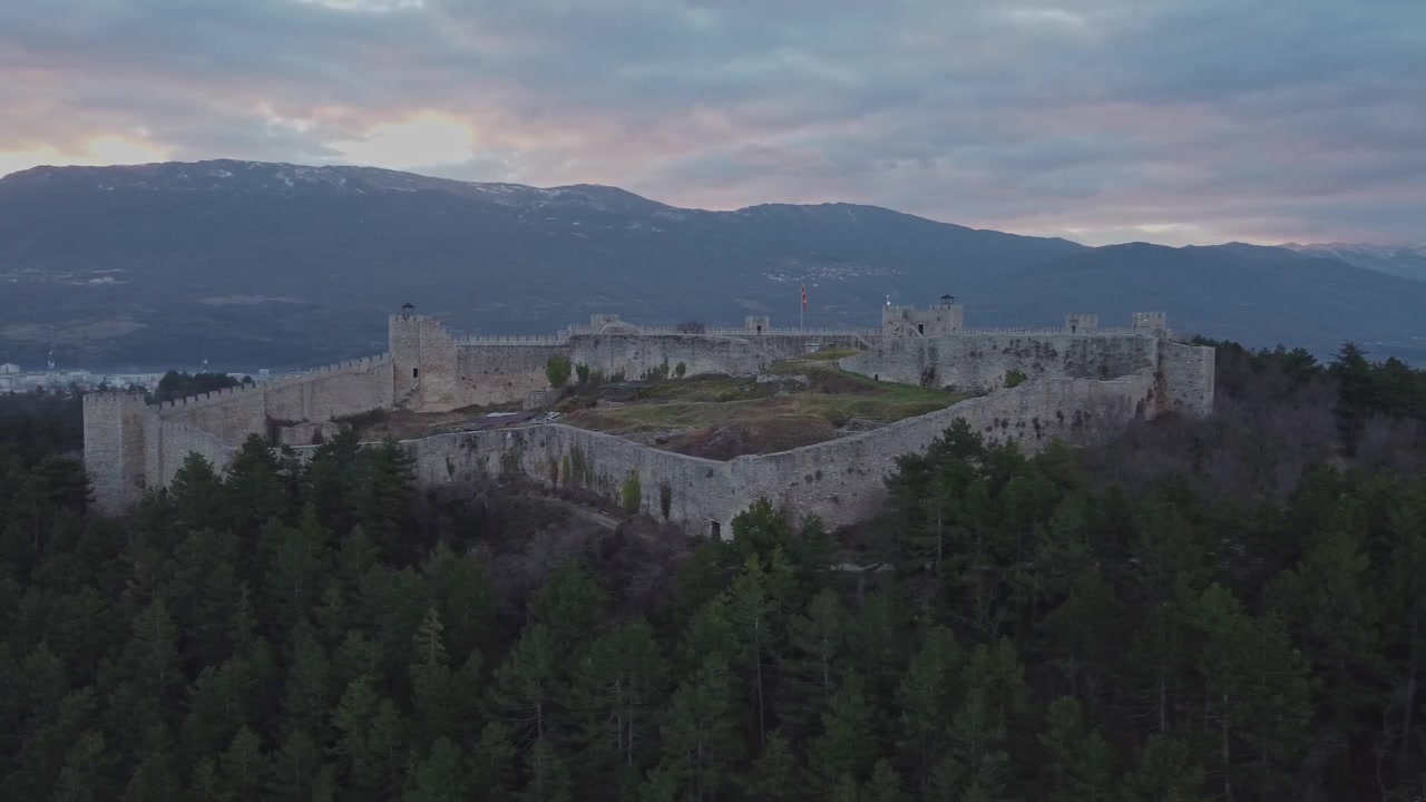 Ampia ripresa aerea della Fortezza di Samuele a Ohrid, Macedonia del Nord, circondata da una pineta verde con una catena montuosa imponente e nuvole colorate al tramonto sullo sfondo.