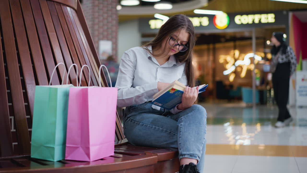 un adulto joven sentado, con las piernas cruzadas, absorto en un libro en el centro comercial, luces bokeh suaves y transeúntes en un fondo borroso añaden una atmósfera vibrante