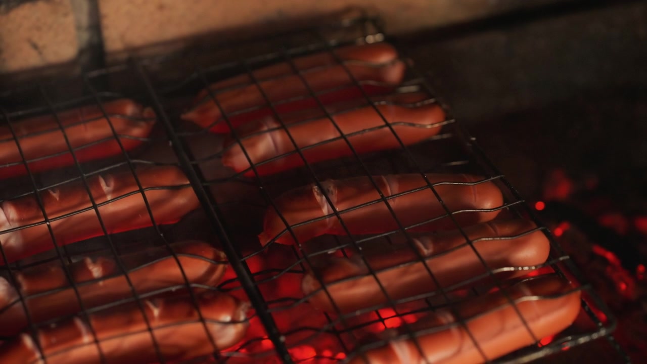 Close low angle view of multiple sausages arranged in parallel on metal grill grate above glowing orange red charcoal embers, with visible sizzling, browning, light charring and warm smoke
