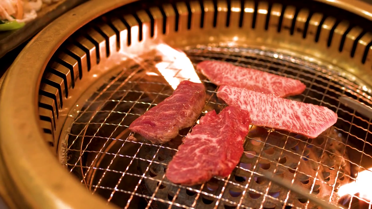 Primo piano di carne di manzo marmorizzata pregiata che cuoce su una griglia barbecue circolare in metallo con fiamme.