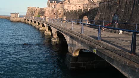 Bridge-to-Castle-Cornet,St-Peter-Port-Guernsey-with-dog-walkers-and-taken-looking-towards-the-castle-and-the-sea-on-sunny-day-with-calm-sea