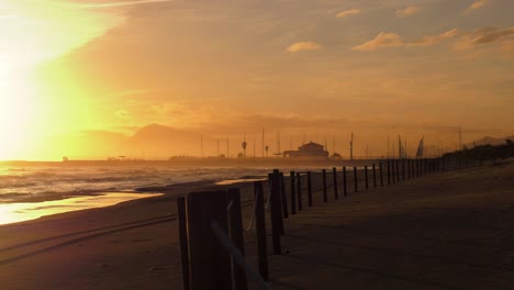 Deserted-sandy-beach-and-dunes-at-sunrise-with-waves-breaking-on-shore,-harbor-in-background