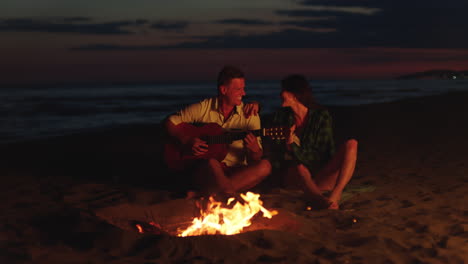 couple playing guitar by campfire on the beach at sunset