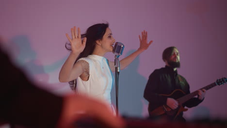 a female singer in a white gown stands in front of a vintage microphone, passionately singing with her hands outstretched. a guitarist plays in the background, and a blurred keyboardist