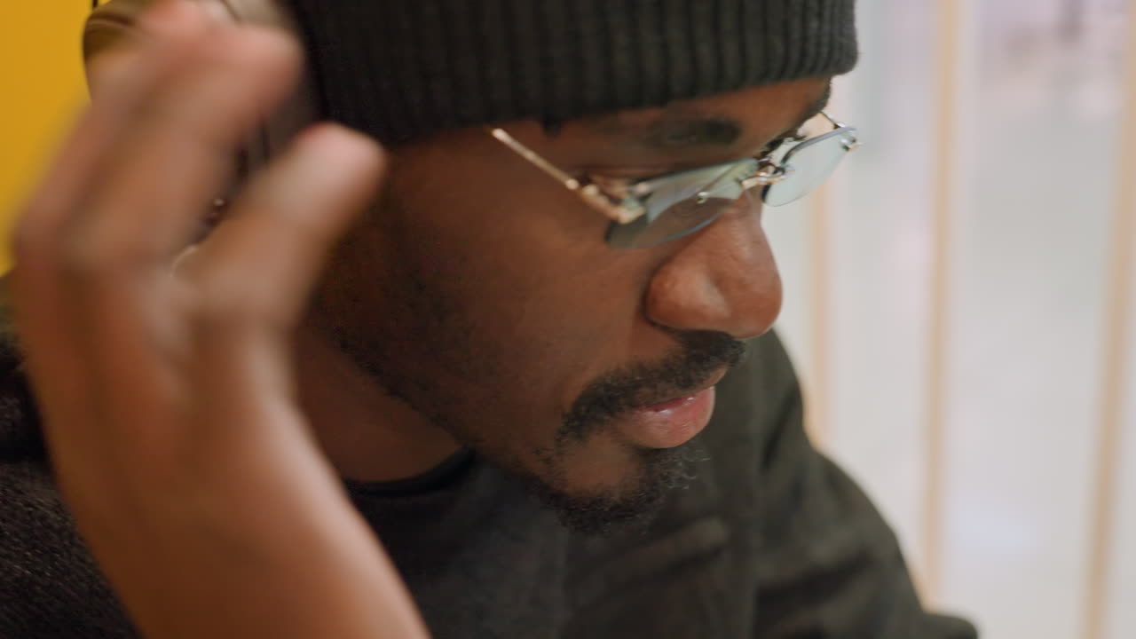 Man inspecting sound with glasses. Intimate profile view of man evaluating audio details. Man wearing glasses and beanie attentively examines music through headphones in cozy cafe setting