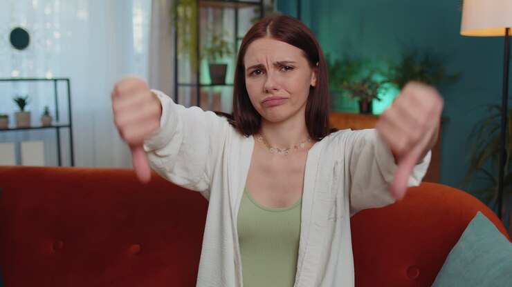 Upset young woman showing thumbs down, dislike bad work, disapproval, dissatisfied feedback at home